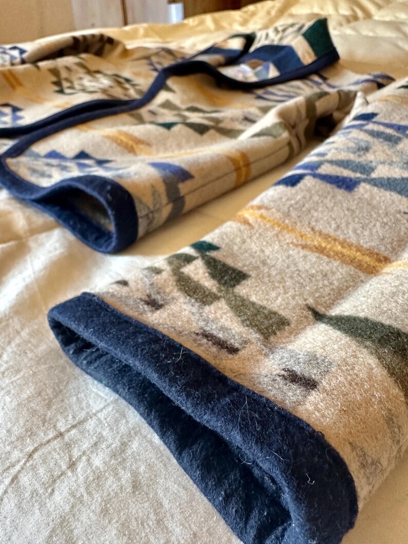 Może przedstawiać: Złożony koc z geometrycznym wzorem w odcieniach beżu, niebieskiego, zieleni i złota. Koc ma ciemnoniebieską obw&oacute;dkę. Wz&oacute;r zawiera tr&oacute;jkąty i kształty przypominające strzały. Koc jest wykonany z miękkiego, teksturowanego materiału.