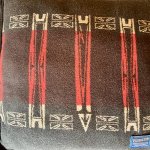 Peut inclure: Un coussin carr&eacute; gris fonc&eacute; avec un motif r&eacute;p&eacute;t&eacute; de rayures verticales rouges et de formes g&eacute;om&eacute;triques blanches et rouges. Le coussin a une bordure noire et une petite &eacute;tiquette bleue avec du texte blanc dans le coin inf&eacute;rieur droit. Le coussin est en tissu tiss&eacute;.