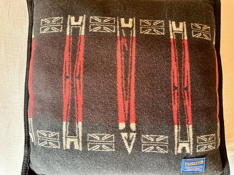 Peut inclure: Un coussin carr&eacute; gris fonc&eacute; avec un motif r&eacute;p&eacute;t&eacute; de rayures verticales rouges et de formes g&eacute;om&eacute;triques blanches et rouges. Le coussin a une bordure noire et une petite &eacute;tiquette bleue avec du texte blanc dans le coin inf&eacute;rieur droit. Le coussin est en tissu tiss&eacute;.