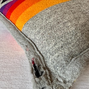 May include: A grey wool cushion with a vibrant rainbow design. The rainbow features bands of red, orange, yellow, purple, and black. The cushion has a grey border and a textured appearance.