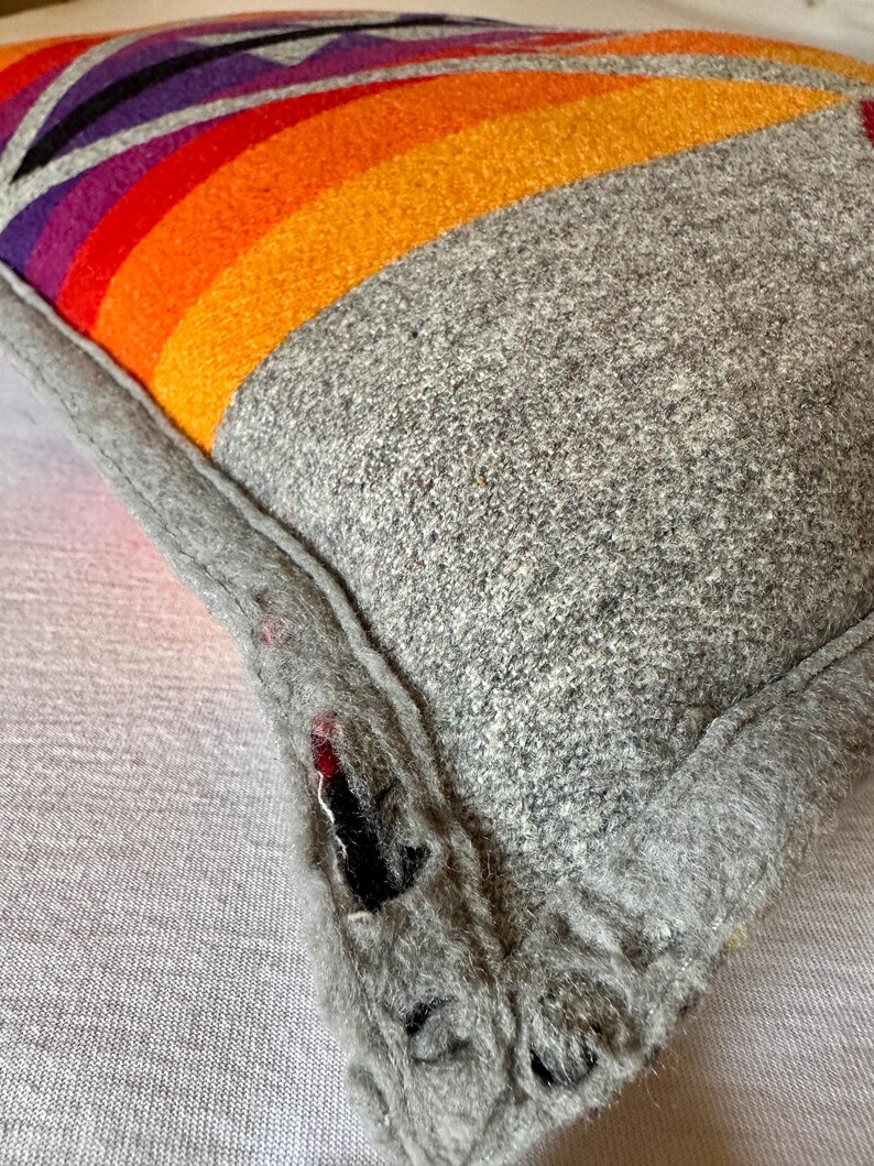 May include: A grey wool cushion with a vibrant rainbow design. The rainbow features bands of red, orange, yellow, purple, and black. The cushion has a grey border and a textured appearance.