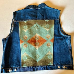 May include: A denim vest with a turquoise, orange, and brown patterned back panel. The vest has a classic collar, button closures, and two front pockets. The vest is made of denim and a woven fabric.