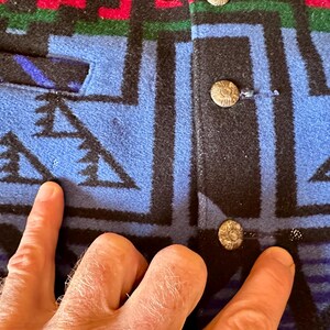 Puede incluir: Primer plano de una chaqueta estampada azul, negra, roja y verde. La chaqueta presenta un dise&ntilde;o geom&eacute;trico con cierres de botones. La tela parece ser lana gruesa o un material similar. La chaqueta tiene un bolsillo en el lado izquierdo.