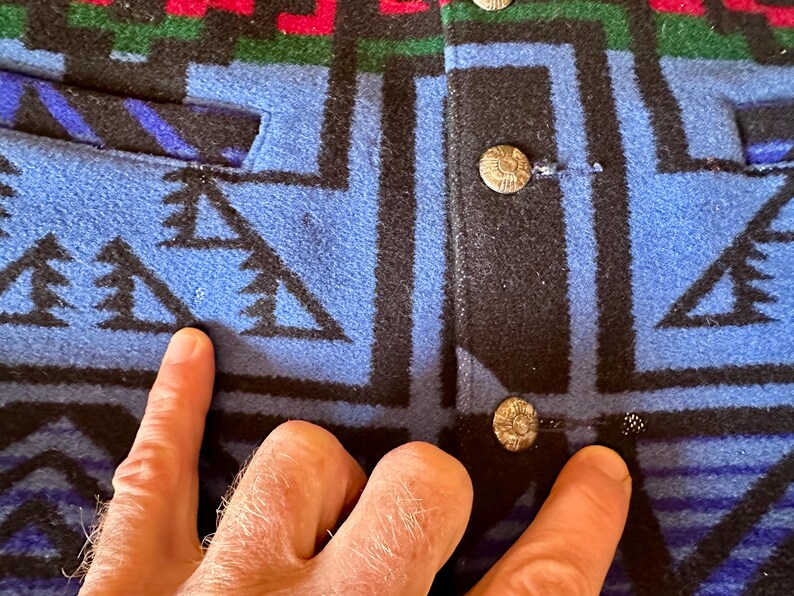 Puede incluir: Primer plano de una chaqueta estampada azul, negra, roja y verde. La chaqueta presenta un dise&ntilde;o geom&eacute;trico con cierres de botones. La tela parece ser lana gruesa o un material similar. La chaqueta tiene un bolsillo en el lado izquierdo.