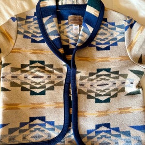 Może przedstawiać: Beżowa wełniana kurtka z geometrycznym wzorem w kolorze niebieskim, zielonym i ż&oacute;łtym. Kurtka ma granatowe wykończenie i kołnierz. Etykieta Pendleton jest widoczna z przodu.