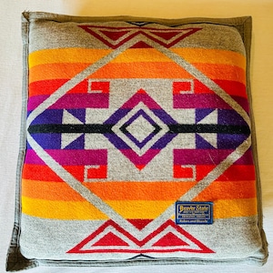 May include: Square cushion with a vibrant geometric design in shades of orange, red, purple, and grey. The cushion features a central diamond pattern and a grey border. A small blue tag with text is visible on the front.