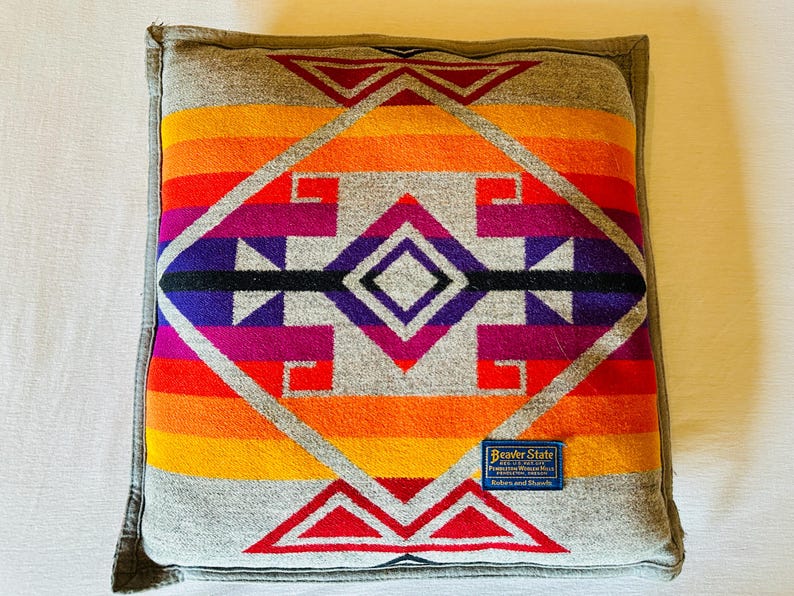May include: Square cushion with a vibrant geometric design in shades of orange, red, purple, and grey. The cushion features a central diamond pattern and a grey border. A small blue tag with text is visible on the front.