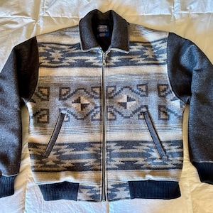 Könnte beinhalten: Eine Jacke mit Reißverschluss und einem Muster im Südwest-Stil in Grau-, Blau- und Cremetönen. Die Jacke hat dunkelgraue Ärmel, einen Kragen sowie gerippte Bündchen und Saum. Das Design umfasst geometrische Formen.