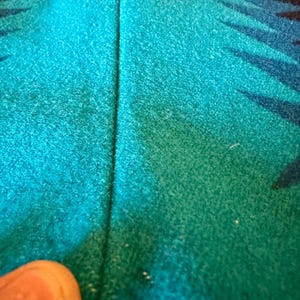 May include: Close-up of a turquoise and teal fabric with a geometric pattern in black and blue. The fabric has a textured appearance and a vertical seam down the center.