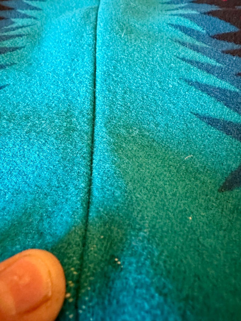 May include: Close-up of a turquoise and teal fabric with a geometric pattern in black and blue. The fabric has a textured appearance and a vertical seam down the center.