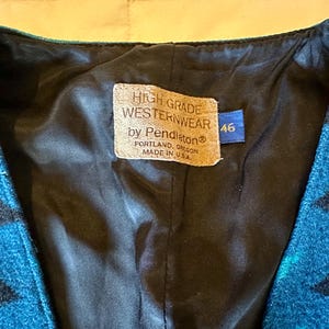 May include: Close-up of a turquoise and black patterned vest with a black lining. The vest has a tag that reads "HIGH GRADE WESTERNWEAR by Pendleton Portland, Oregon MADE IN U.S.A." A blue tag with the number "46" is also visible.