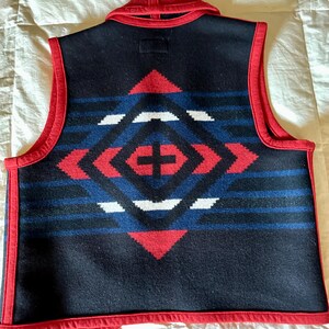 May include: A navy blue vest with a geometric pattern in red, white, and blue. The vest has a red collar and trim. The back of the vest features a diamond-shaped design with a cross in the center.