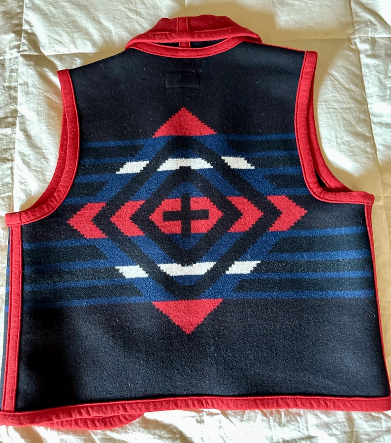 May include: A navy blue vest with a geometric pattern in red, white, and blue. The vest has a red collar and trim. The back of the vest features a diamond-shaped design with a cross in the center.