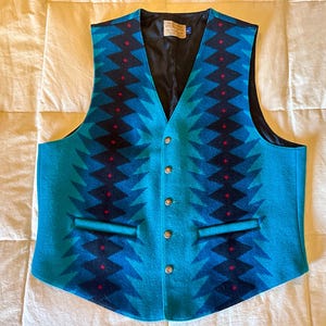May include: A turquoise vest with a geometric pattern in shades of blue and black, accented with small red details. The vest has a V-neck, a black lining, and four silver buttons down the front. It features two front pockets.