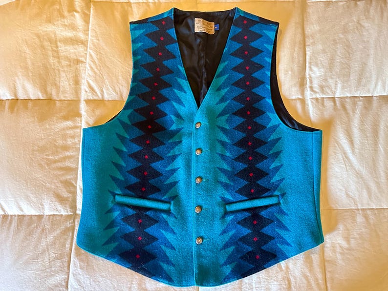 May include: A turquoise vest with a geometric pattern in shades of blue and black, accented with small red details. The vest has a V-neck, a black lining, and four silver buttons down the front. It features two front pockets.