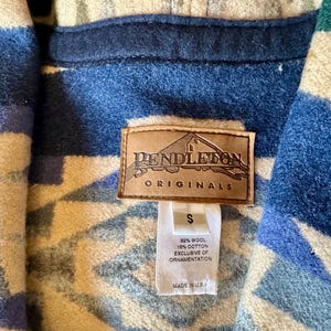 Może przedstawiać: Zbliżenie kurtki Pendleton Originals. Kurtka ma geometryczny wz&oacute;r w odcieniach niebieskiego, kremowego i białego. Widoczna jest brązowa sk&oacute;rzana metka z logo Pendleton, a także metka wskazująca rozmiar S i skład tkaniny.