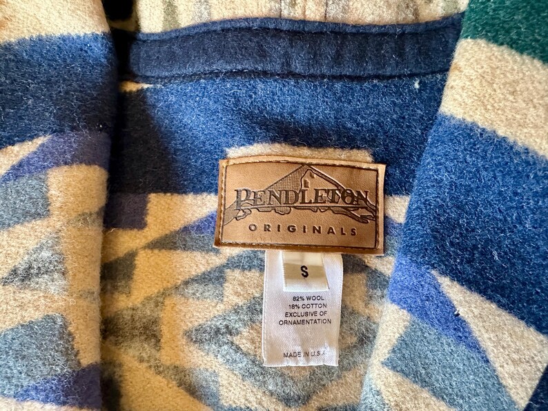 Może przedstawiać: Zbliżenie kurtki Pendleton Originals. Kurtka ma geometryczny wz&oacute;r w odcieniach niebieskiego, kremowego i białego. Widoczna jest brązowa sk&oacute;rzana metka z logo Pendleton, a także metka wskazująca rozmiar S i skład tkaniny.