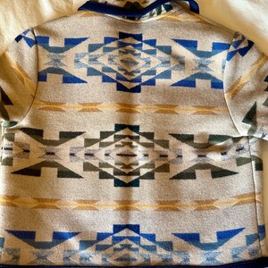 Może przedstawiać: Beżowa kurtka z geometrycznym wzorem w odcieniach niebieskiego, zielonego i ż&oacute;łtego. Kurtka ma granatowy kołnierz i wykończenia. Symetryczny wz&oacute;r pokrywa cały tył kurtki.