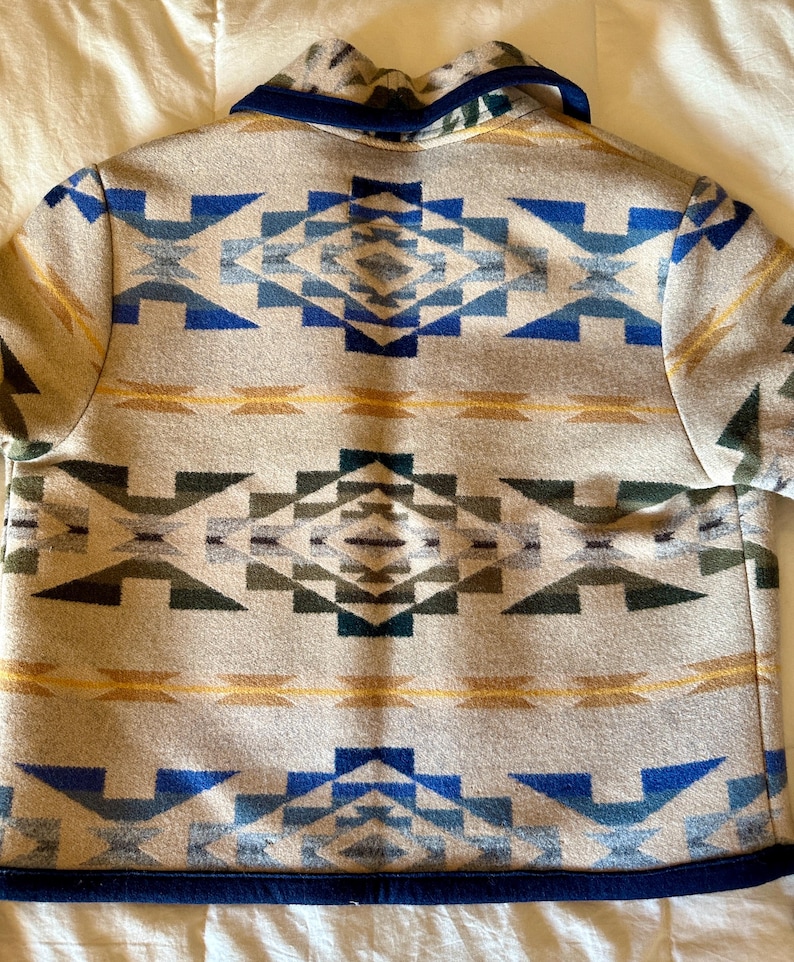 Może przedstawiać: Beżowa kurtka z geometrycznym wzorem w odcieniach niebieskiego, zielonego i ż&oacute;łtego. Kurtka ma granatowy kołnierz i wykończenia. Symetryczny wz&oacute;r pokrywa cały tył kurtki.