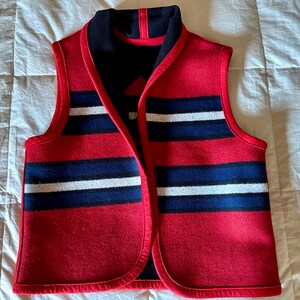 May include: A red wool vest with horizontal stripes in navy blue and white. The vest has a navy blue collar and trim. The vest is sleeveless and has a V-neck design. The vest is made of a thick wool material.