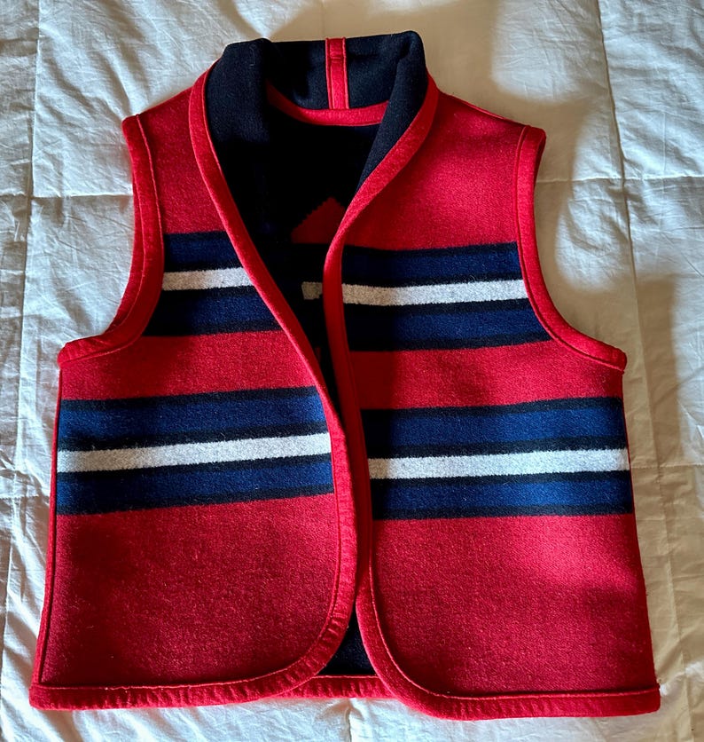 May include: A red wool vest with horizontal stripes in navy blue and white. The vest has a navy blue collar and trim. The vest is sleeveless and has a V-neck design. The vest is made of a thick wool material.