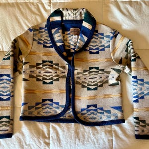 May include: A beige jacket with a geometric pattern in blue, green, and yellow. The jacket has a shawl collar, a navy blue trim, and long sleeves. The jacket is made of a thick, woven material.