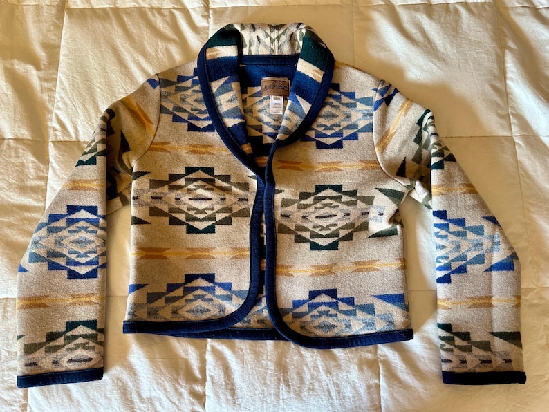 Może przedstawiać: Beżowa kurtka z geometrycznym wzorem w kolorze niebieskim, zielonym i ż&oacute;łtym. Kurtka ma kołnierz szalowy, granatowe wykończenie i długie rękawy. Wykonana z grubego, tkanego materiału.