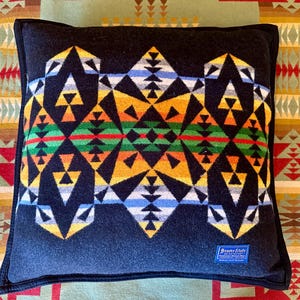 May include: A square, dark blue pillow with a geometric pattern in yellow, orange, green, red, white, and blue. The pillow has a black border and a small tag in the lower right corner. The pillow is on a patterned blanket.