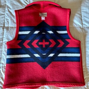 May include: Red wool vest with a geometric pattern in navy blue, black, and white. The vest features a shawl collar and a tag at the back. The pattern is centered on the back of the vest.