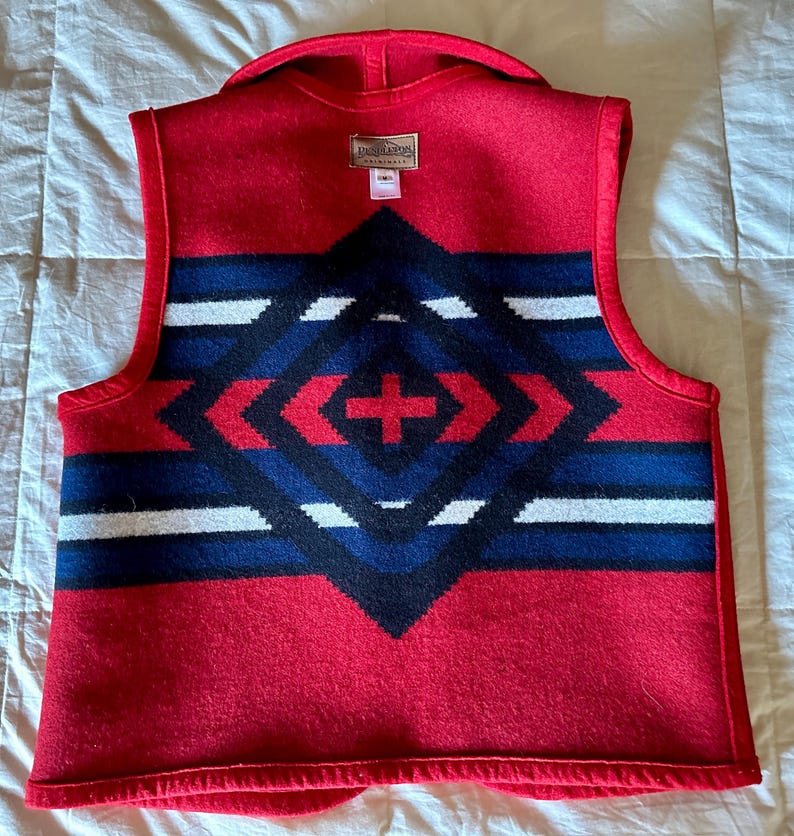 May include: Red wool vest with a geometric pattern in navy blue, black, and white. The vest features a shawl collar and a tag at the back. The pattern is centered on the back of the vest.