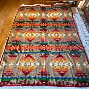 Puede incluir: Una manta grande y rectangular con un patrón geométrico en tonos marrón, naranja, rojo, verde y azul. El diseño presenta formas de diamantes y triángulos. La manta tiene un borde rojo y un reverso blanco.