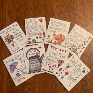 May include: A collection of eight hand-drawn cards with inspirational quotes and floral illustrations. Each card features unique artwork, including a watering can, a beach scene, and a turkey. The cards are arranged on a wooden surface.