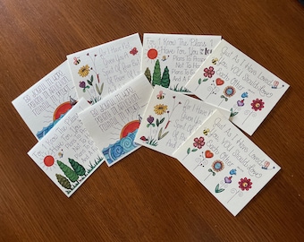 Op de afbeelding: Een verzameling kleurrijke, handgeschilderde kaarten met inspirerende citaten en bloemmotieven. De kaarten zijn in verschillende kleuren, waaronder rood, blauw, groen, geel en roze. De citaten op de kaarten zijn: "Wees blij in hoop, geduldig in tegenspoed, trouw in gebed", "Want ik weet welke plannen ik voor jullie heb", "Want ik heb jullie geen geest van angst gegeven, maar een geest van kracht, liefde en bezonnenheid", en "Zoals ik jullie heb liefgehad, zo moeten jullie elkaar liefhebben".