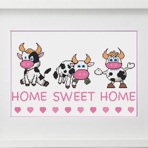 May include: Framed cross-stitch art featuring three cartoon cows with black and white spots, brown horns, and pink accents. The phrase "HOME SWEET HOME" is stitched in pink below the cows, with pink hearts. The artwork is in a white frame.