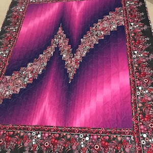 May include: A quilt with a gradient pattern of pink and purple. The quilt is made with a patchwork design and has a black border with a pattern of red and silver circles.