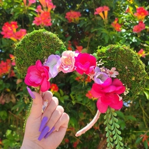 May include: A floral headband with moss-covered ears, adorned with vibrant pink, purple, and white flowers. The headband is held up by a hand with long purple nails, set against a backdrop of green foliage and red flowers.