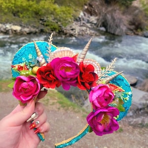 May include: A pair of turquoise blue and gold mouse ears with pink and red fabric flowers, seashells, and a gold horn accent.