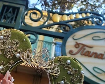 Può includere: Orecchie di Minnie Mouse in velluto verde con ricami dorati, perle bianche e una tiara scintillante. Le orecchie sono davanti a un cartello che dice "Tiana's".