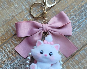 May include: A white cat keychain with a pink bow and a pearl-beaded circle. The cat has pink ears and cheeks. A large pink bow and gold-colored keyring complete the design. The background is a wooden surface.