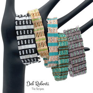 Puede incluir: Cinco pulseras de cuentas coloridas con un patrón de rayas. Las pulseras se muestran en un soporte negro. El texto "Deb Roberti Tila Stripes" es visible debajo de las pulseras.