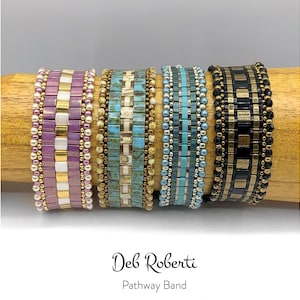 May include: Four beaded bracelets in shades of purple, turquoise, and black, with gold and silver accents. The bracelets are displayed on a wooden dowel. The text "Deb Roberti Pathway Band" is below the bracelets.