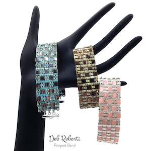 May include: Three beaded bracelets with a geometric pattern. The bracelets are turquoise, brown, and pink. The bracelets are on a black hand-shaped display stand. The text "Deb Roberti Parquet Band" is below the bracelets.