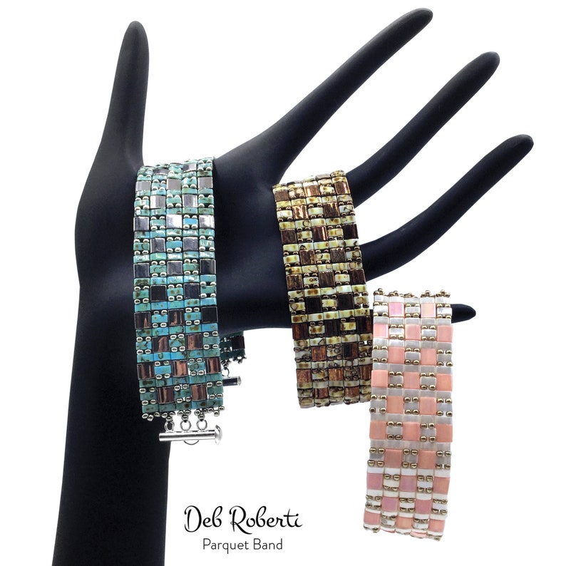 May include: Three beaded bracelets with a geometric pattern. The bracelets are turquoise, brown, and pink. The bracelets are on a black hand-shaped display stand. The text "Deb Roberti Parquet Band" is below the bracelets.