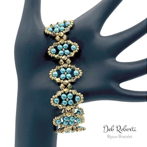 Puede incluir: Un brazalete de cuentas turquesa y dorado con un diseño geométrico. El brazalete está en una mano de maniquí negra. "Deb Roberti Bijoux Bracelet" está escrito debajo del brazalete.