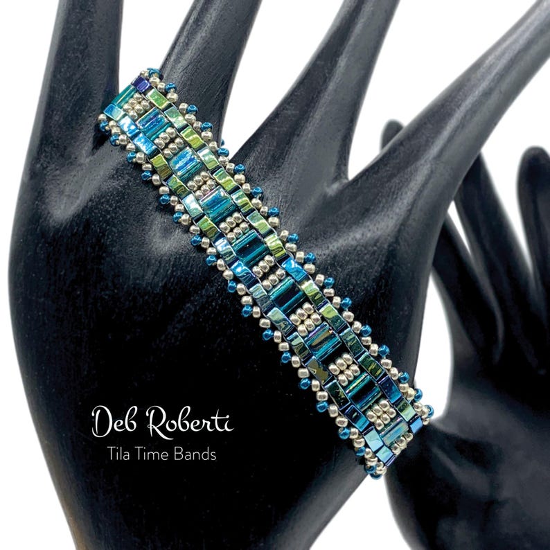 May include: A blue, green, and silver beaded bracelet with a geometric pattern. The bracelet is on a black mannequin hand. The text "Deb Roberti Tila Time Bands" is below the bracelet.