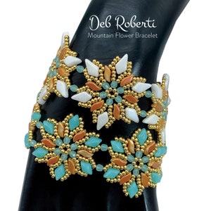May include: A beaded bracelet featuring a design of orange, white and turquoise flowers. The bracelet is displayed on a black mannequin arm. The text "Deb Roberti Mountain Flower Bracelet" is above the bracelet.
