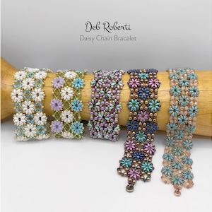 以下が含まれることがあります： 白、緑、紫、青、桃色など、さまざまな色のビーズで作られたデイジーチェーンブレスレットが5つ。白い背景に、木製のダボにブレスレットが展示されています。「Deb Roberti Daisy Chain Bracelet」というテキストが上部にあります。
