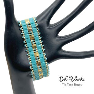 May include: A turquoise and gold beaded bracelet on a black hand model. The bracelet is made with small square beads and has a geometric pattern. The text "Deb Roberti Tila Time Bands" is visible below the bracelet.