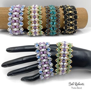 May include: Four beaded bracelets in various colors, including lavender, green, teal, and black, are displayed on a wooden cylinder and a black hand. The bracelets feature intricate beadwork with pearls and crystals. The text "Deb Roberti Thalia Band" is visible.