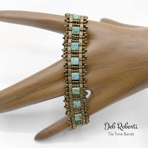 May include: A beaded bracelet with a gold and turquoise color scheme. The bracelet is made with small, square beads and has a delicate, intricate design. The bracelet is on a wooden hand model. The text "Deb Roberti Tila Time Bands" is visible below the bracelet.